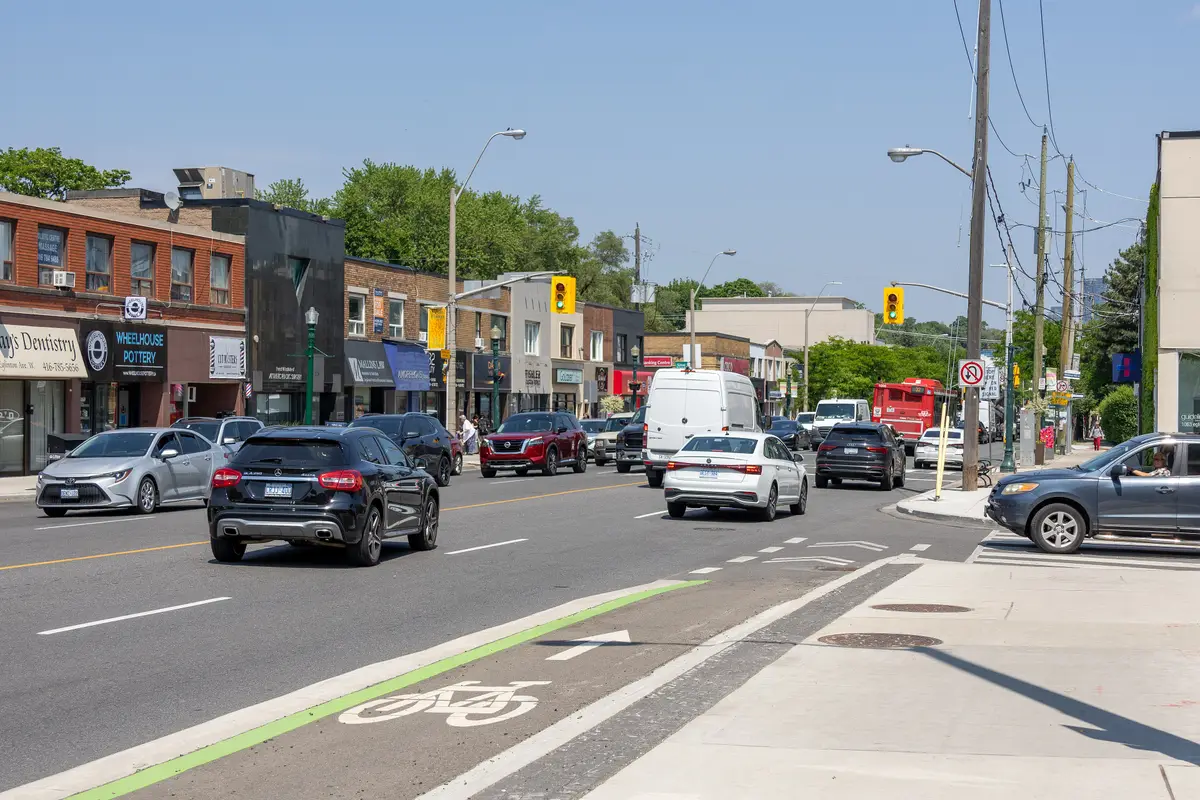 Eglinton Avenue West on a sunny summer day, with shopfronts along the street and a painted bike lane at the curb
