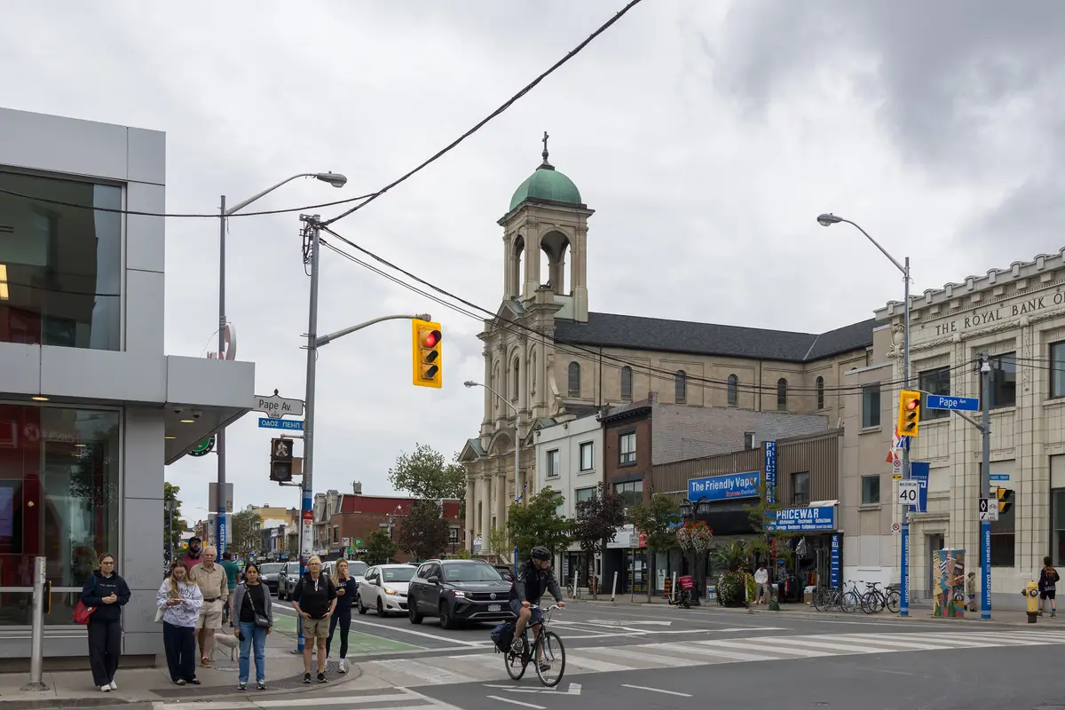 Danforth Avenue at Pape Avenue in Greektown, with a green-domed church, shopfronts, pedestrians waiting at the corner and a cyclist on the road