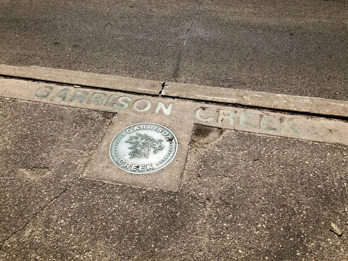 A round metal medallion reading GARRISON CREEK, set in a concrete sidewalk beside a curb, with the words GARRISON CREEK stamped into the curb above it