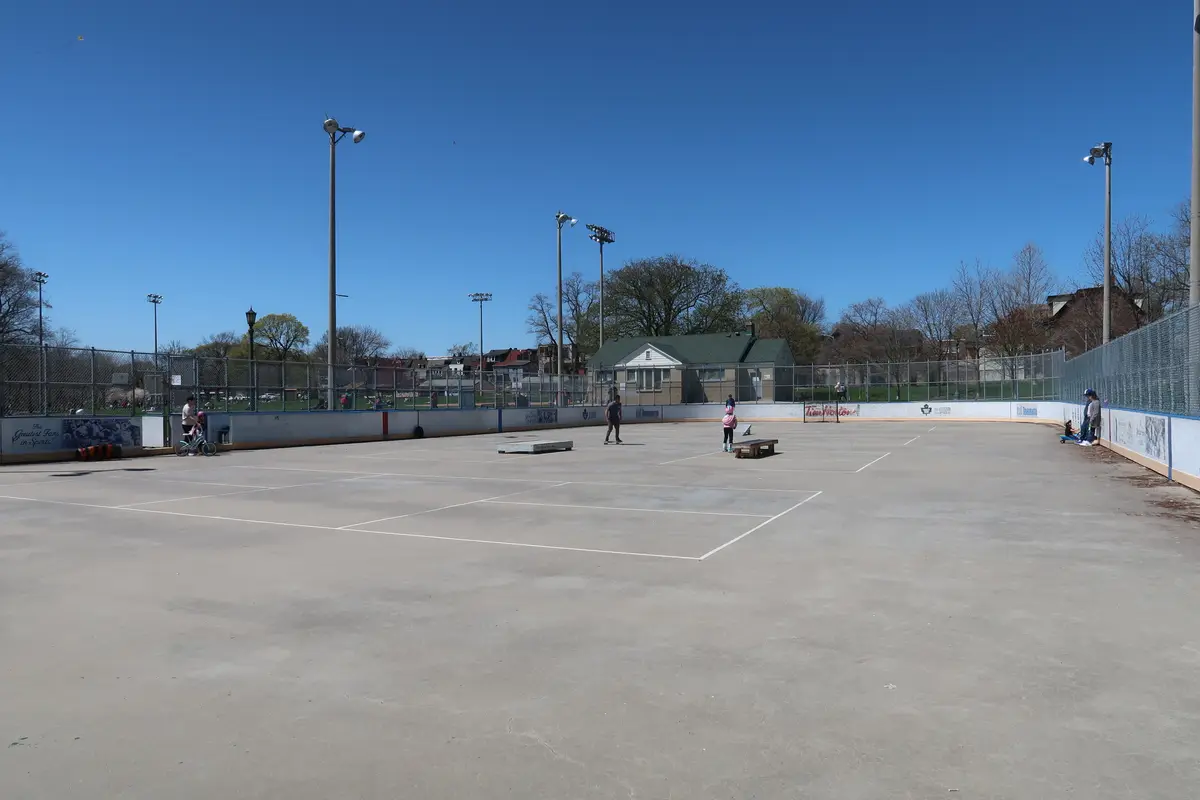 Outdoor concrete sports court with white painted lines, a high board fence and tall floodlight poles under a clear blue sky, with a few people playing
