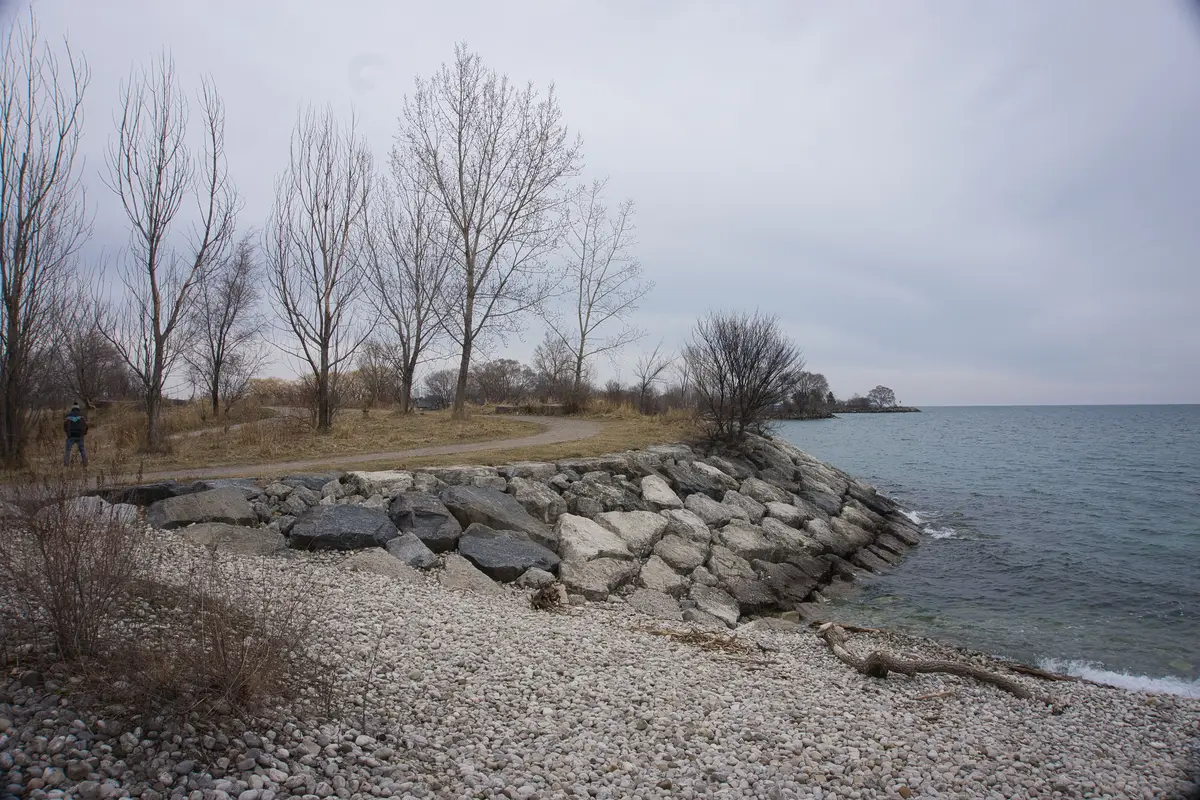 A gravel path beside a cobble beach and rock shoreline at Mimico Waterfront Park on a grey day, with bare trees and Lake Ontario to the right