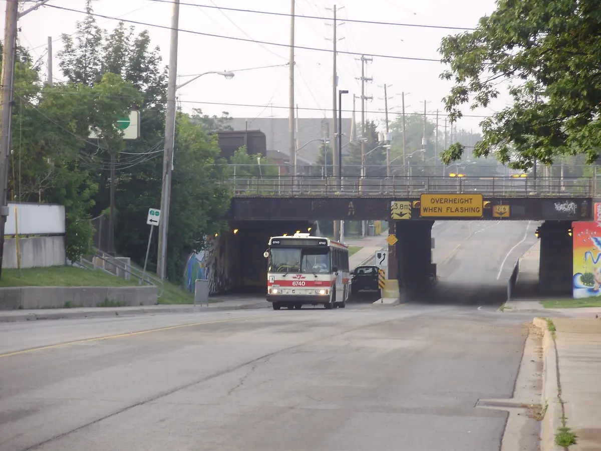 A TTC bus emerging from beneath the railway bridge on Royal York Road at Judson Street in Mimico, with trees and murals along the underpass