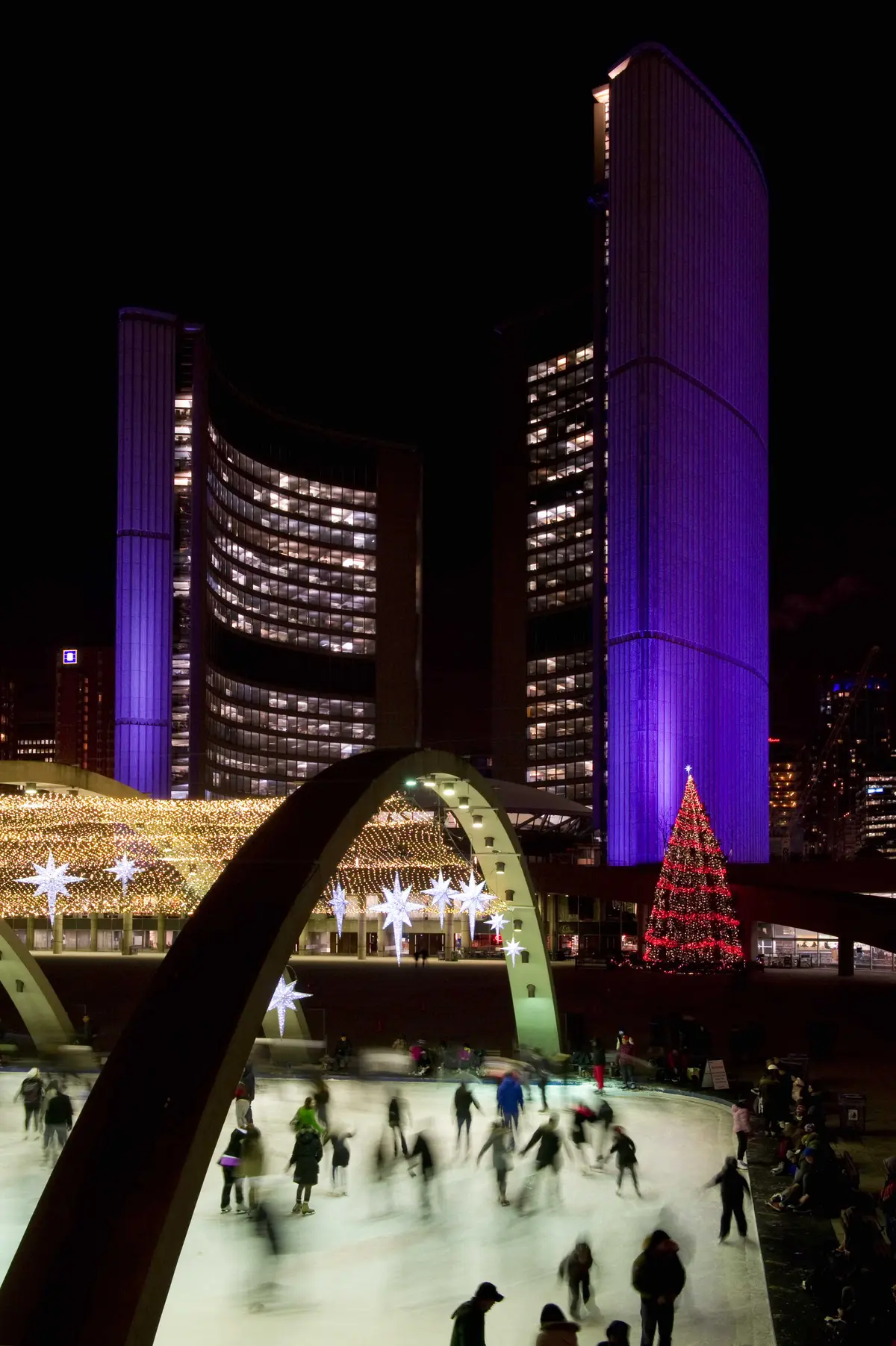 Skaters circling the rink at night, with illuminated towers behind the ice.