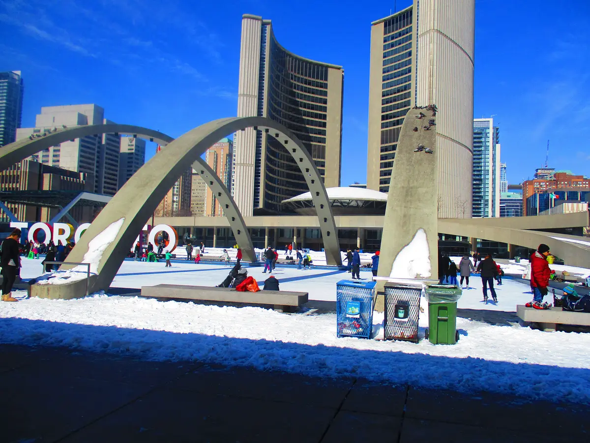 Skaters on the Nathan Phillips Square rink under a blue sky, with the white arches and City Hall towers beside the ice