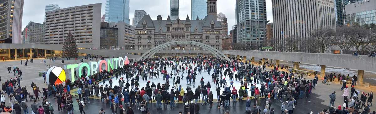 A wide panorama of the Nathan Phillips Square rink, with skaters, arches and City Hall in the background
