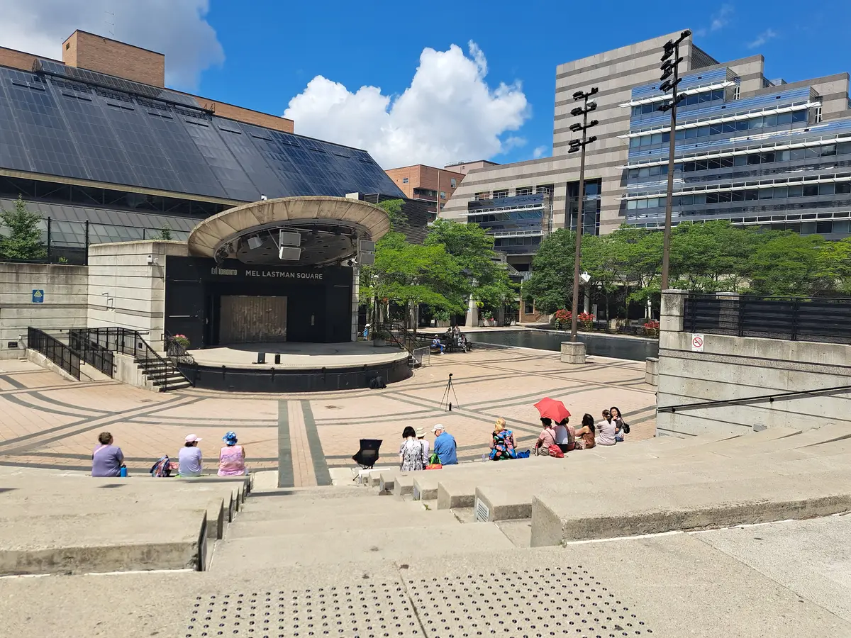 Stepped seating and the round stage of the amphitheatre at Mel Lastman Square on a sunny day, with a few people sitting on the lower level