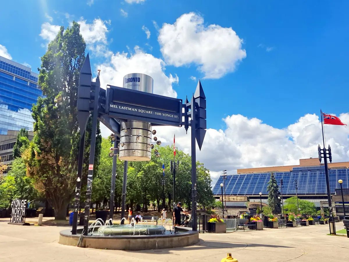 The fountain and street sign at the entrance to Mel Lastman Square under a blue sky, with office towers behind
