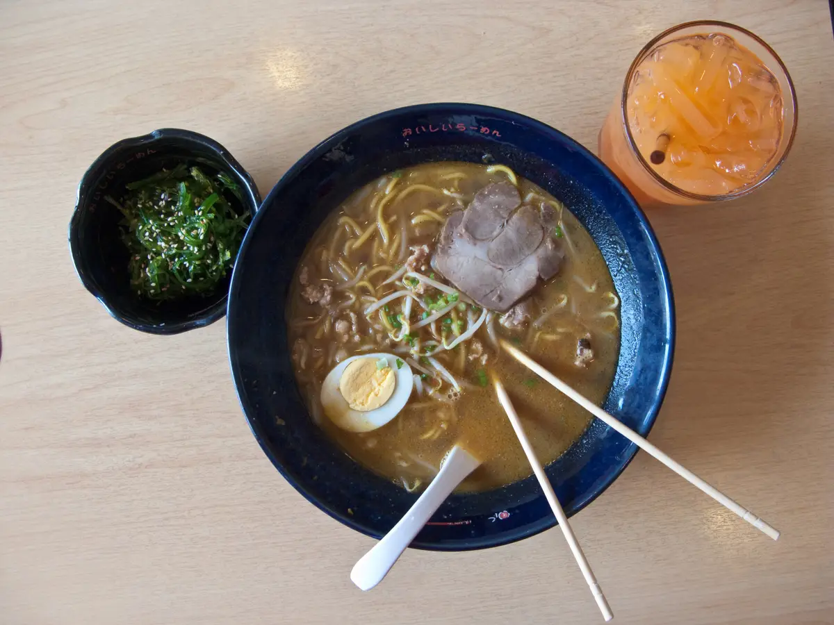 A bowl of shoyu ramen with sliced pork, a halved egg and noodles, beside a small dish of greens