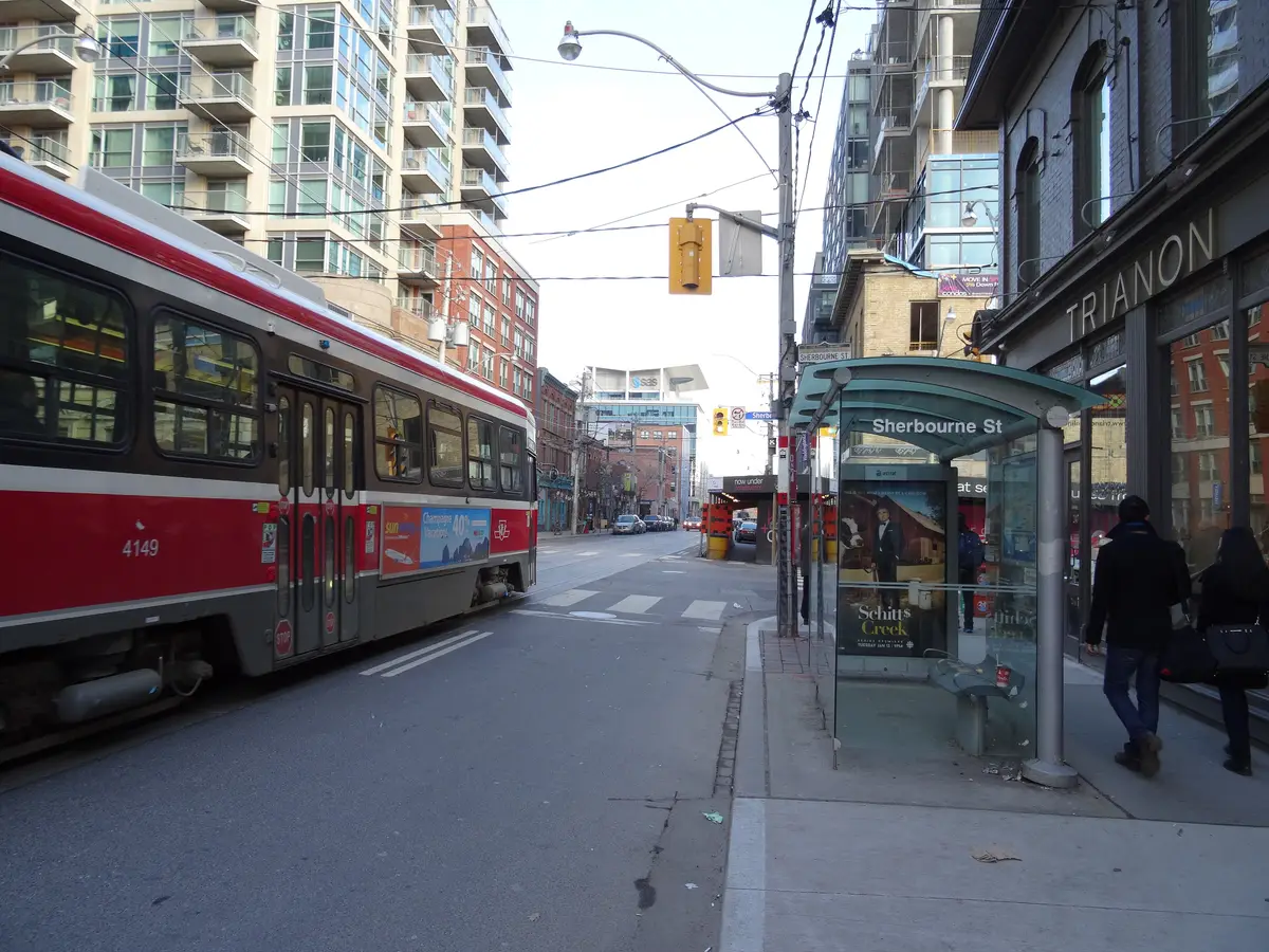A red TTC streetcar passing a stop on King Street with shops and apartment buildings along the block