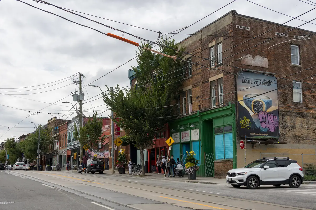 Brick storefronts and a green-trimmed corner building along Queen Street West in Parkdale, with streetcar wires overhead