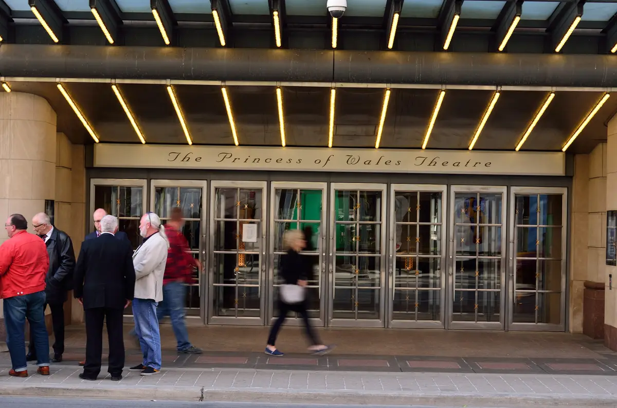 The entrance doors of the Princess of Wales Theatre, with the theatre's name on the sign above and people passing on the sidewalk