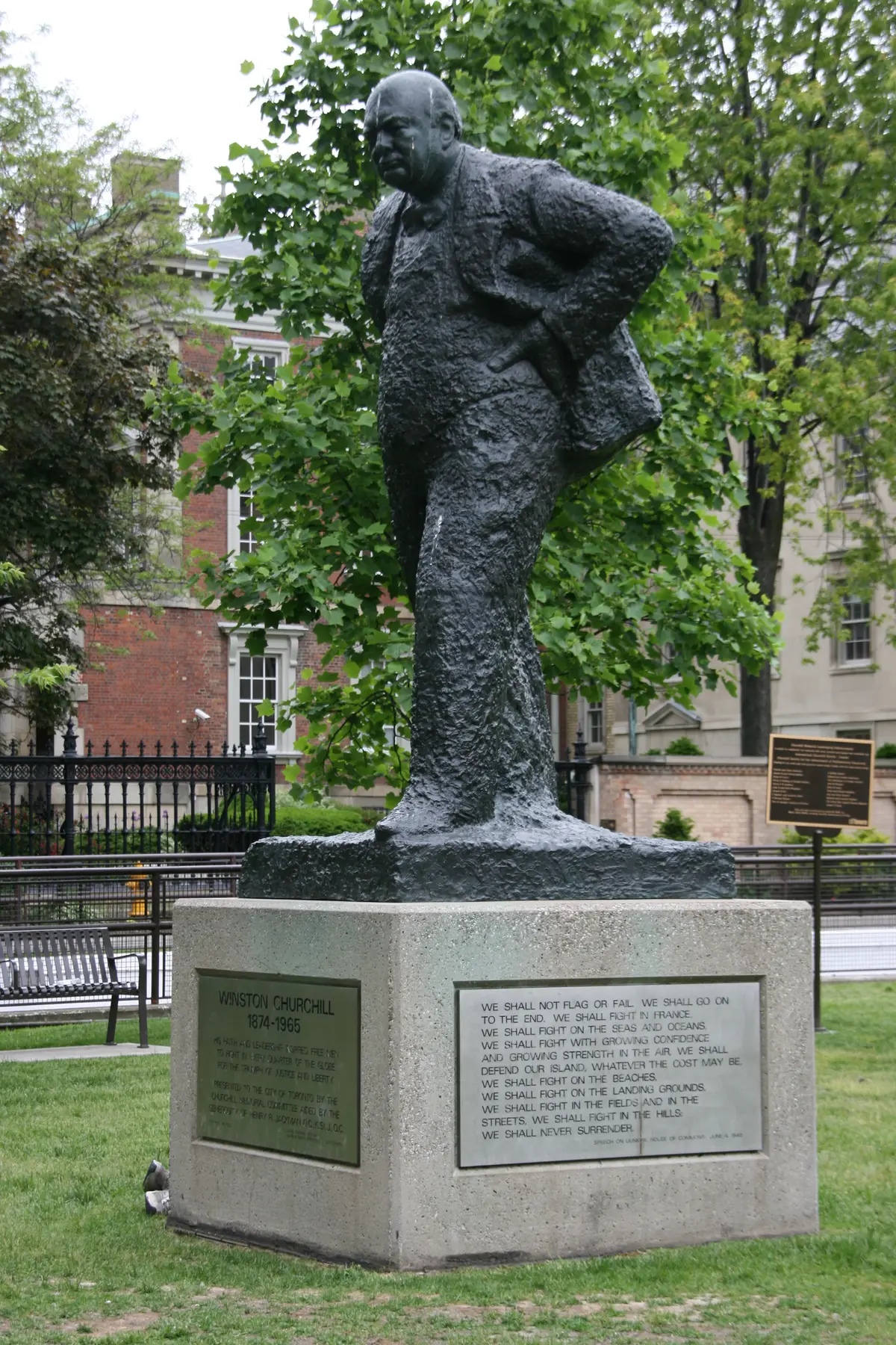 A dark, rough-textured statue of Winston Churchill on a stone plinth, with trees and brick buildings behind