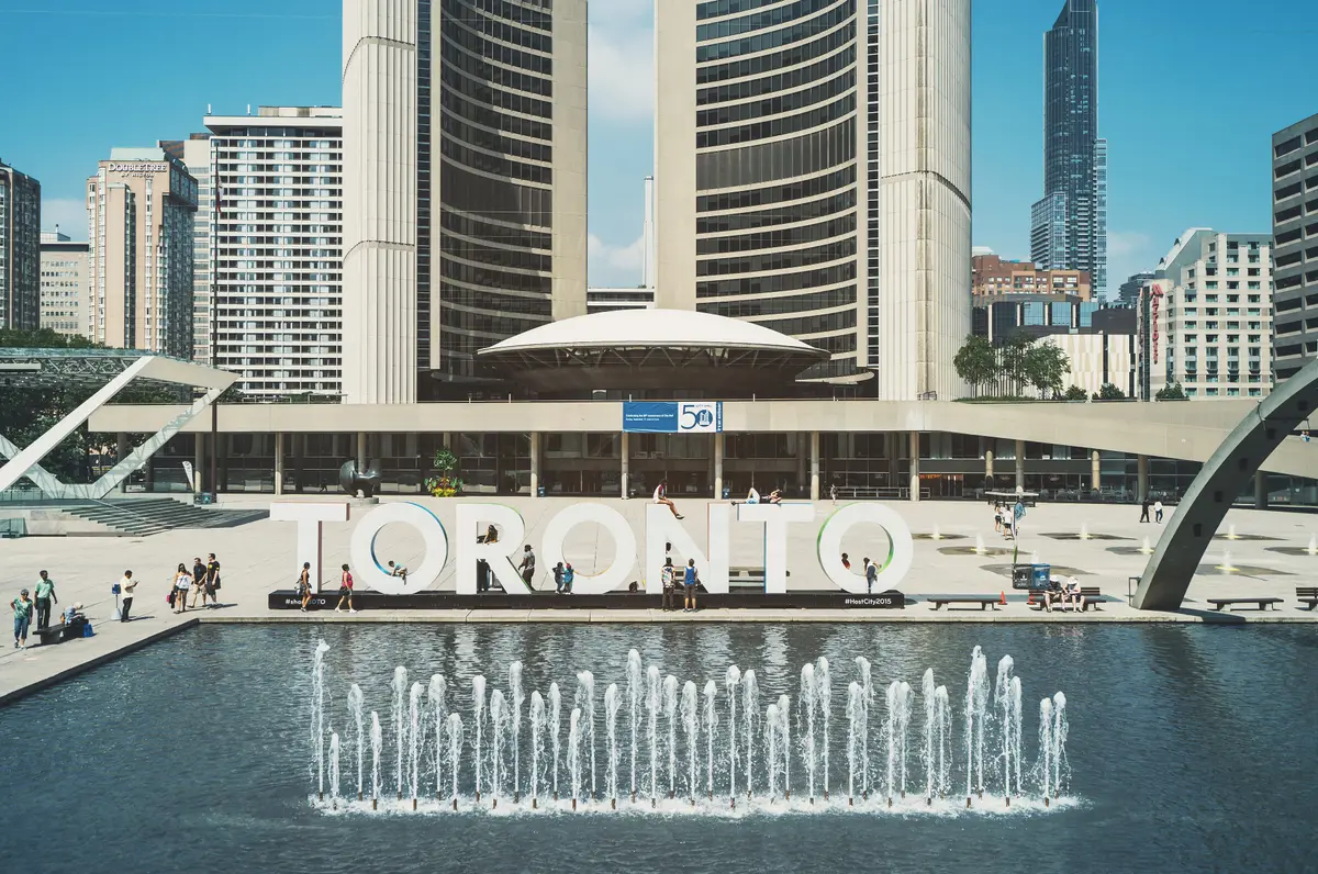 Nathan Phillips Square on a sunny day, with large TORONTO letters above a fountain pool, people walking and curved towers behind