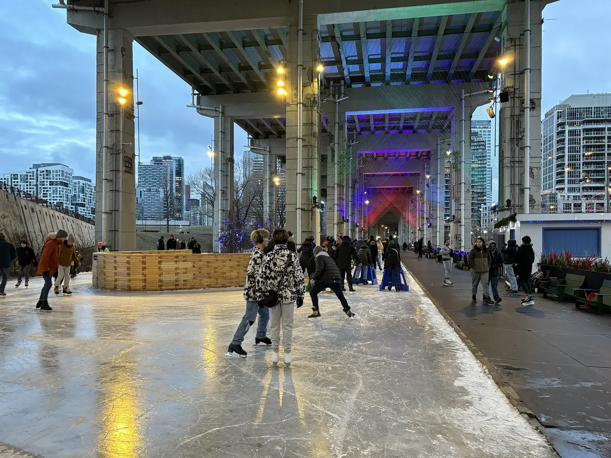 Skaters glide along the Bentway ice trail beneath the raised Gardiner Expressway, with coloured lights glowing under the concrete columns.