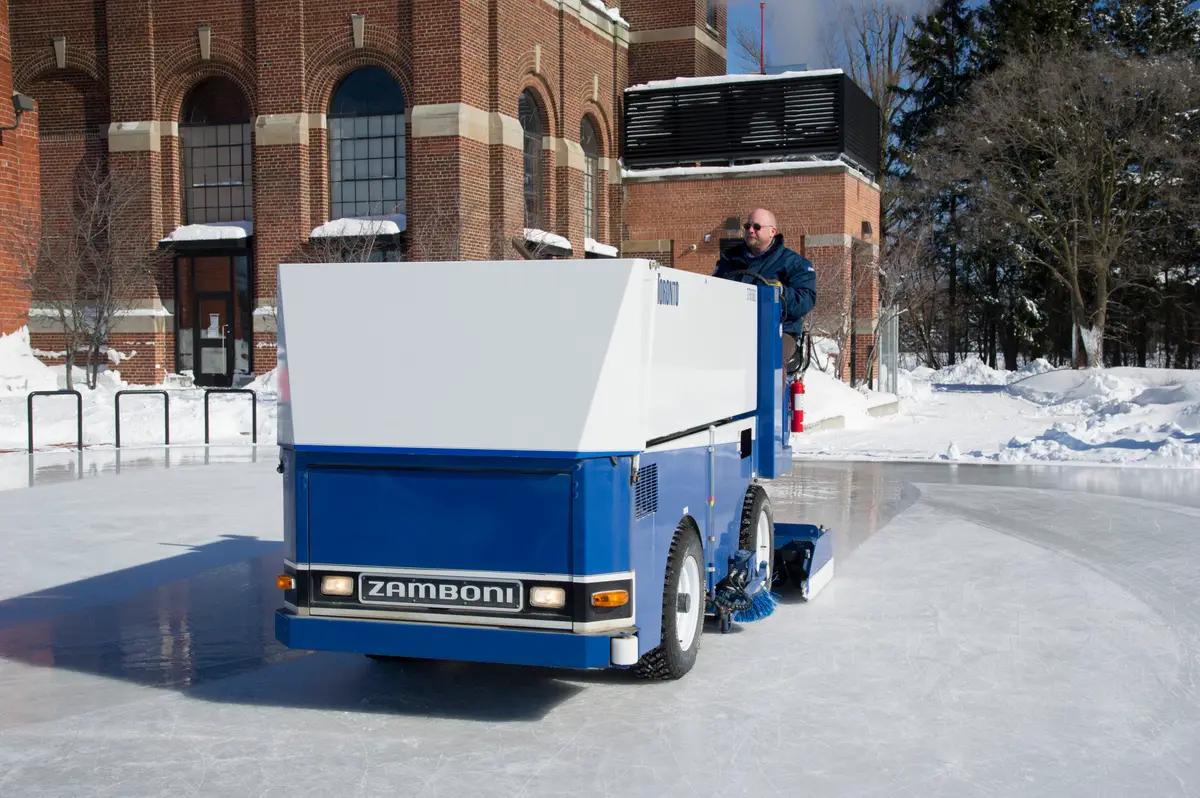 A blue Zamboni resurfacing the ice beside snowbanks and a brick building at Colonel Samuel Smith Park.