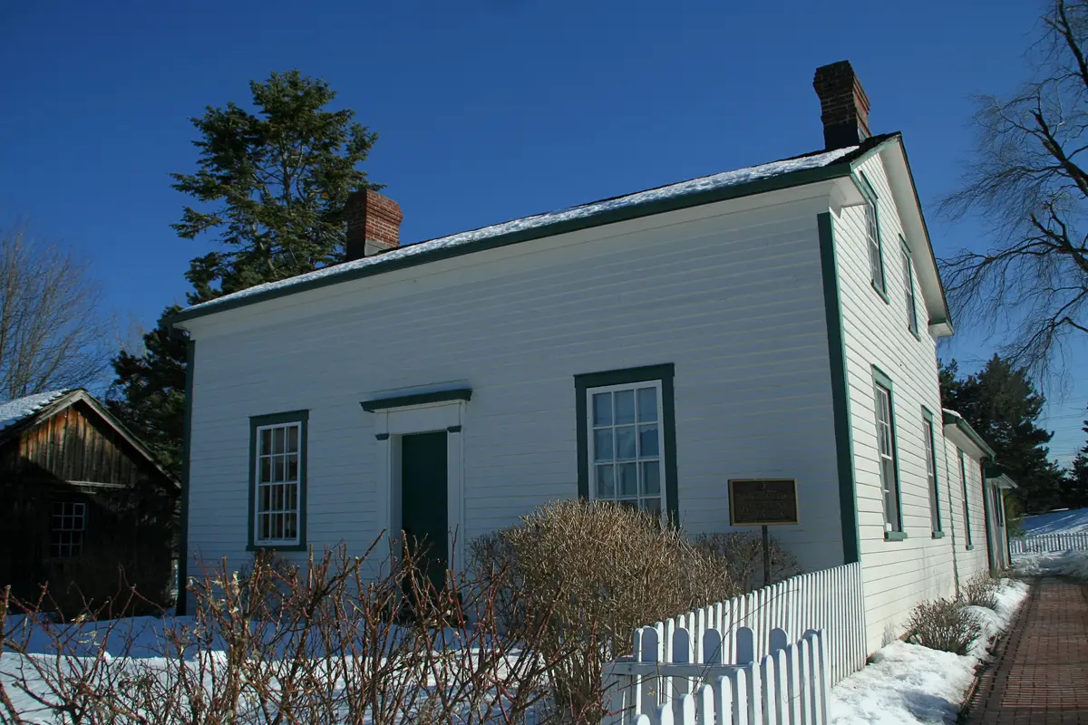 A white clapboard house with green trim and a brick chimney, with snow on the roof, a white picket fence and a brick path.