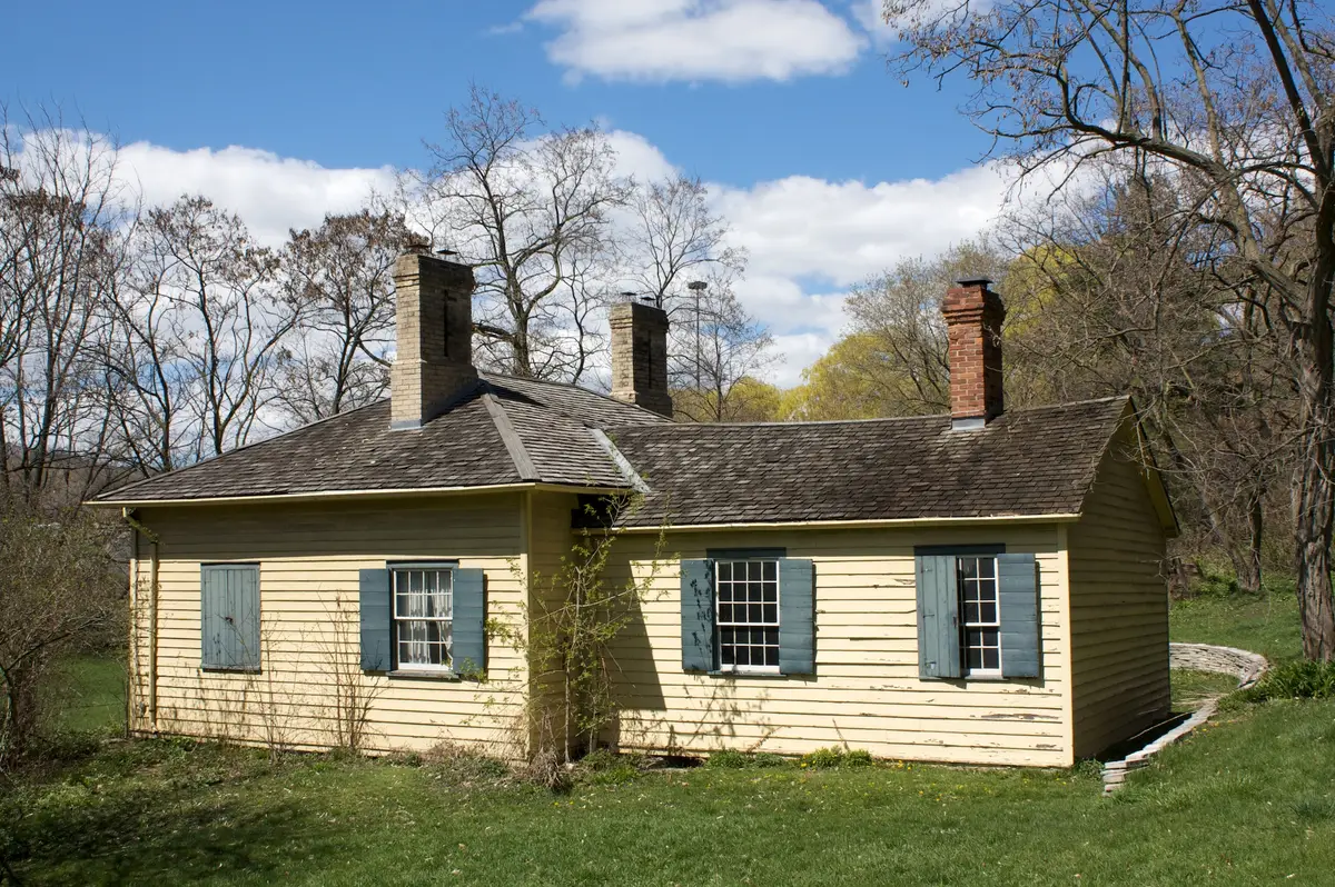A pale yellow clapboard house with blue-grey shutters and three chimneys, set among bare spring trees and green lawn.