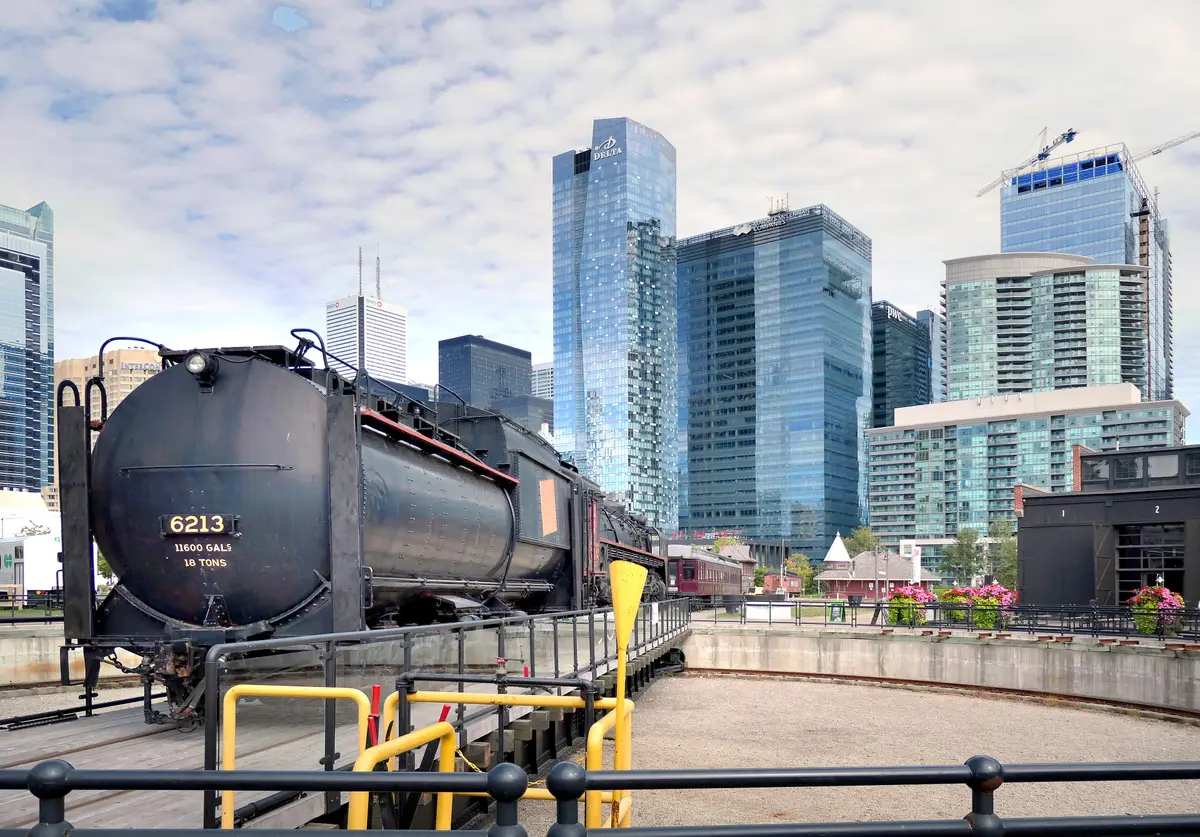 A black rail tank car on a turntable at Roundhouse Park with glass towers of downtown rising behind.