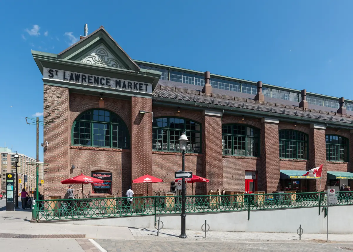 Brick facade of the St. Lawrence Market building with its lettered gable, arched windows and a patio under red umbrellas on a sunny day