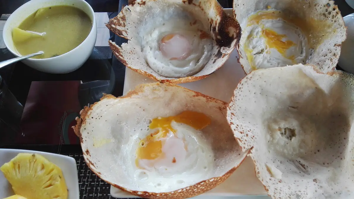 Four bowl-shaped hoppers, three with an egg cooked in the centre, on a tray beside a small bowl of soup.