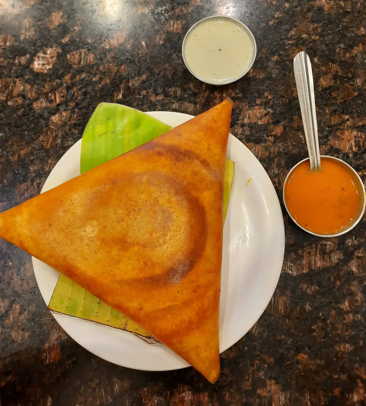 A folded golden dosa on a plate with a small bowl of coconut chutney and a bowl of sambar.