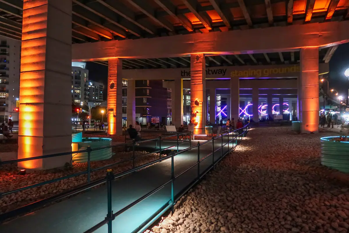 Orange-lit concrete columns under the Gardiner Expressway at night, with a lit path beside pebbled ground