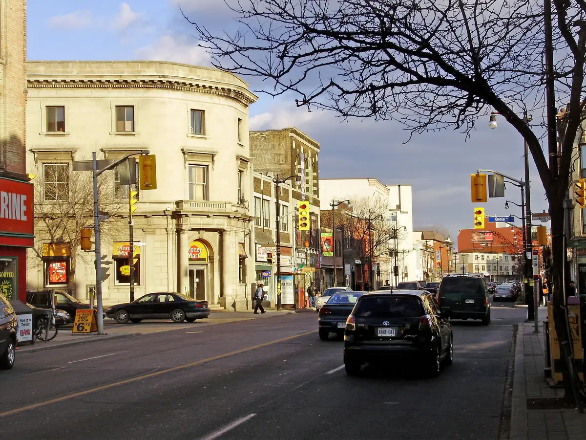 A cream-coloured heritage bank building on the corner and a row of shops along Dundas Street West at Keele Street, looking east