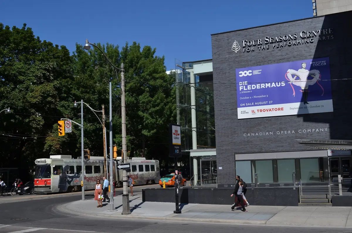 A streetcar passes the dark stone and glass corner of the Four Seasons Centre on a sunny day
