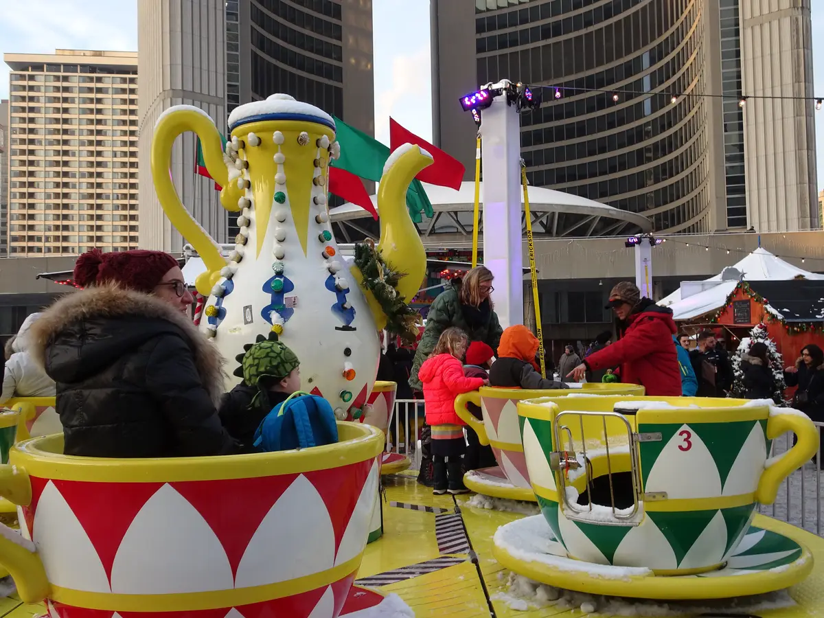 Riders on a teacup ride with a holiday market and the City Hall towers behind