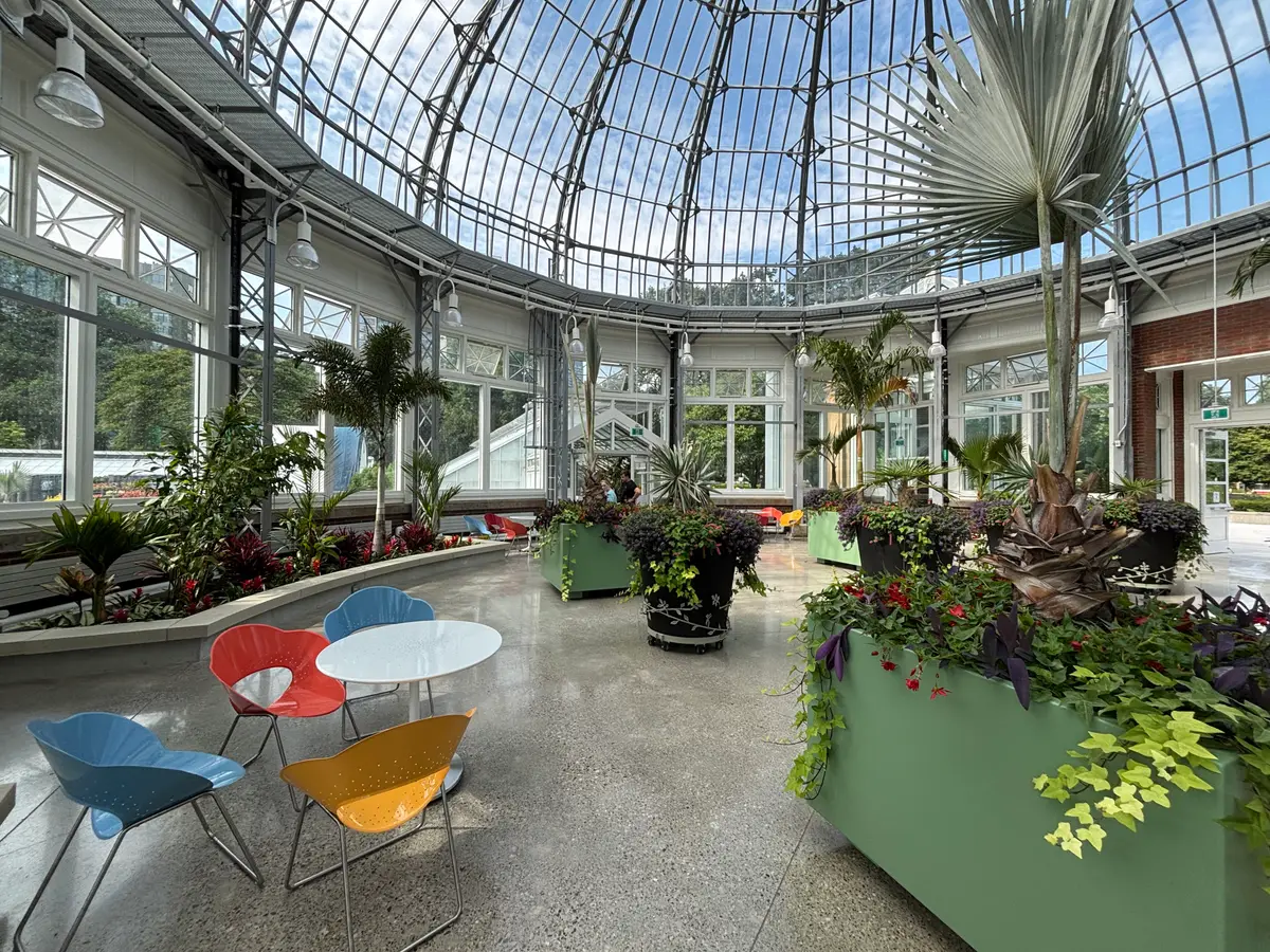 The sunlit glass-domed interior of the Allan Gardens Conservatory, with planters, palm leaves and café chairs.