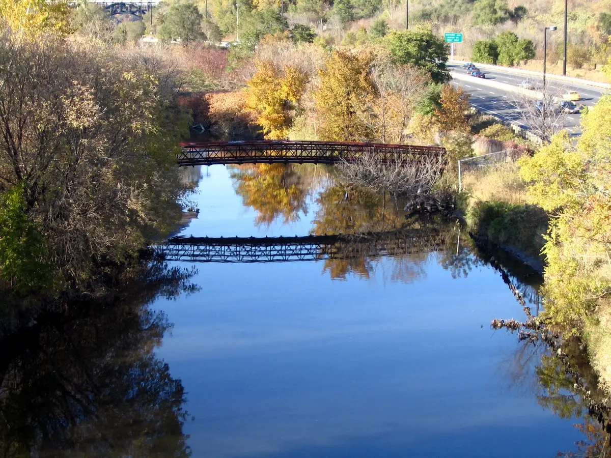 The Don River reflecting blue sky and autumn-coloured trees, with a rail footbridge crossing the water and the Don Valley Parkway at right.