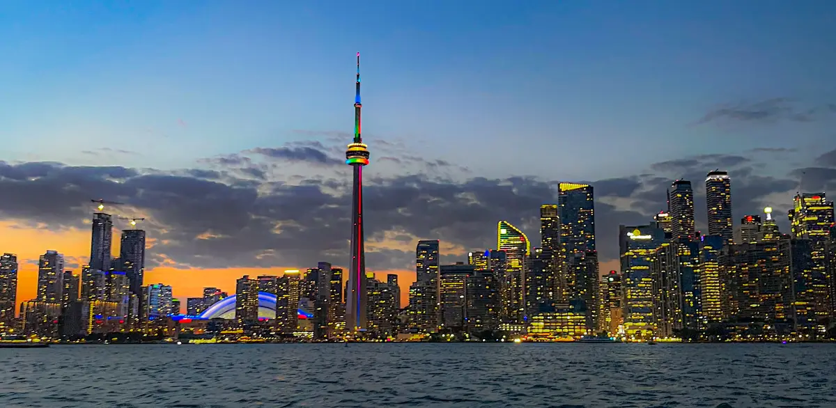 The CN Tower lit in rainbow colours above the Toronto skyline at dusk, seen across the water