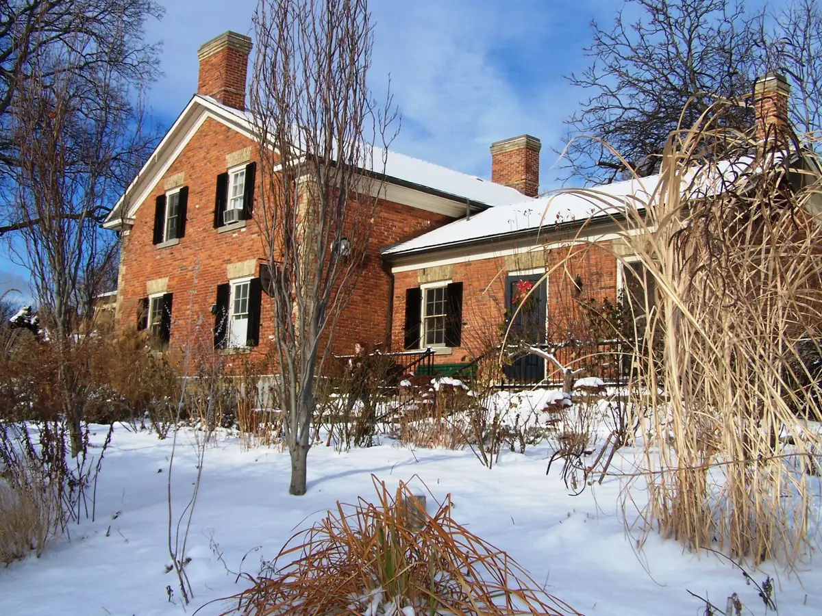 A red brick farm building with black shutters beside snowy gardens