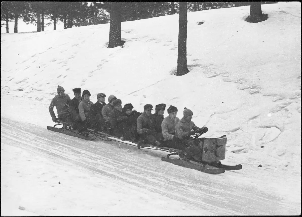 A group of riders in winter coats and caps seated along a long sled with a steering wheel, sliding down a snowy slope, in an archive photo