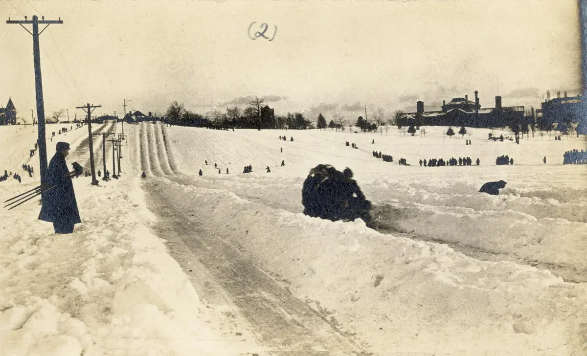 Sledders and onlookers on a snowy hill at Riverdale Park in a photograph from 1900, with a few buildings along the horizon