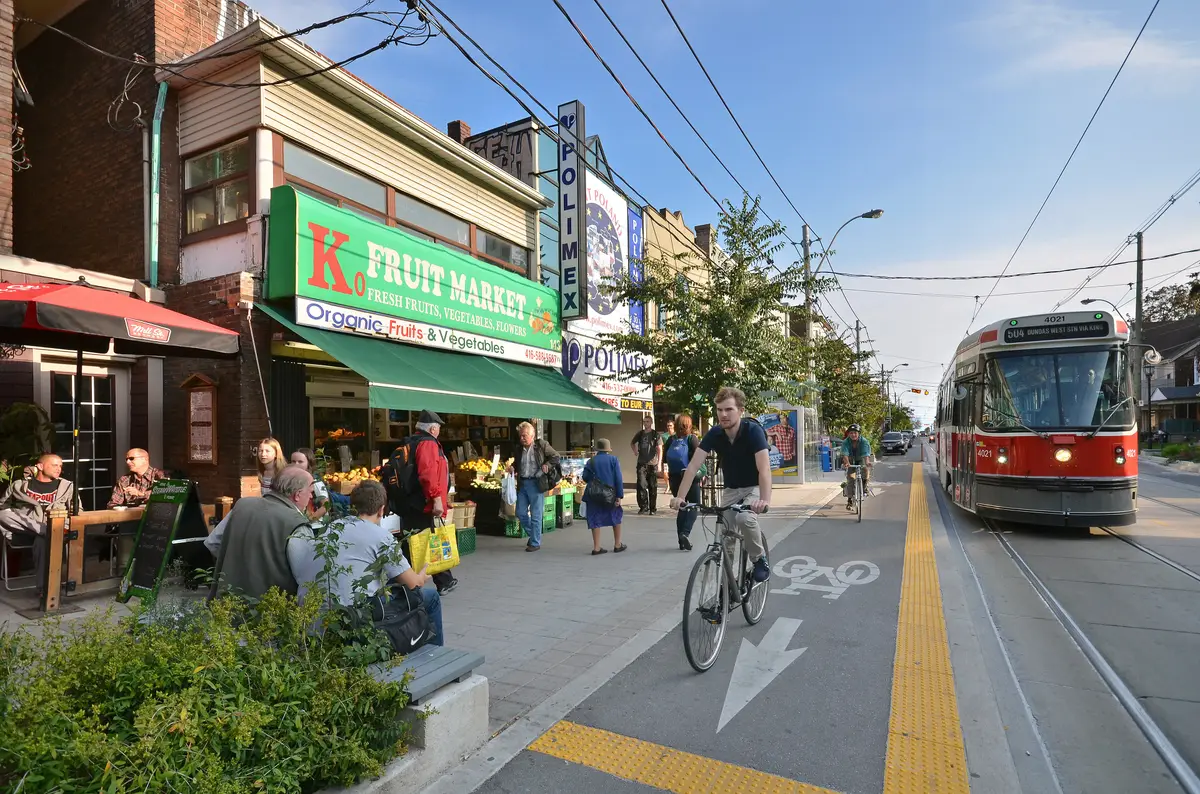 A sunny Roncesvalles Avenue scene with a fruit and vegetable stand, cyclists and a red streetcar