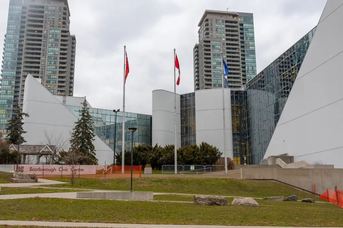 The angular white and glass Scarborough Civic Centre with flagpoles and condo towers behind.