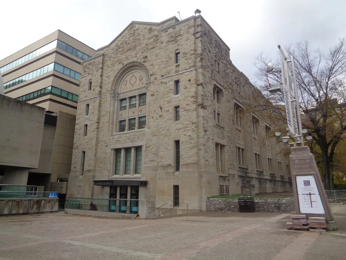 The stone Royal Ontario Museum building with arched windows, seen across a paved plaza on a grey day, with a bare tree and an ROM sign at the right