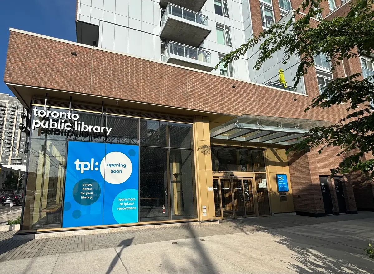 The glass front of the Junction Triangle branch of Toronto Public Library, with a blue banner in the window and a tree at the entrance