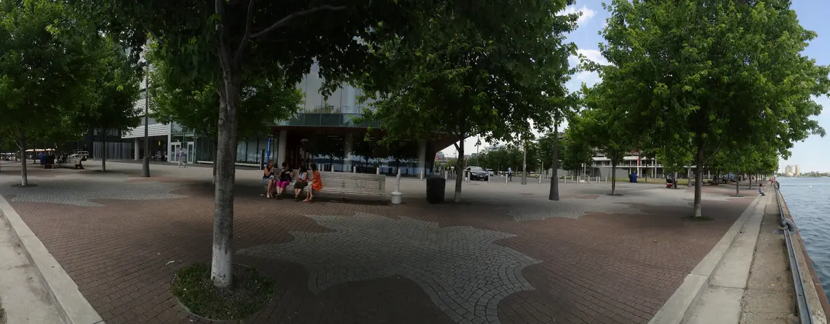 A paved promenade beside Sherbourne Common, lined with leafy trees and benches along the water.