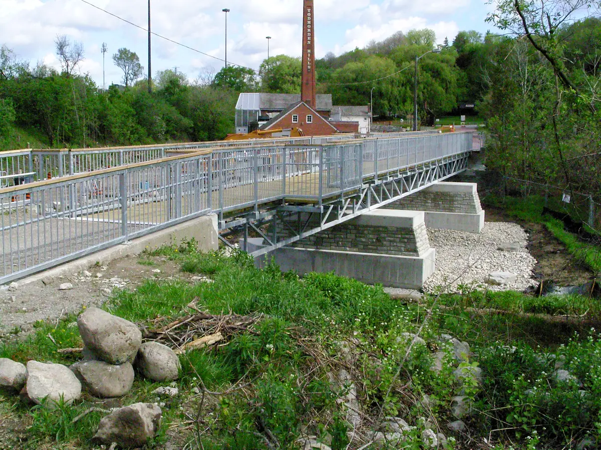 A metal footbridge with railings crosses a gravel riverbed, with a brick chimney marked TODMORDEN MILLS behind it