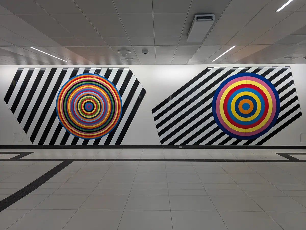 Concentric coloured circles set against black and white diagonal stripes on a white station wall, with a tiled floor in front.