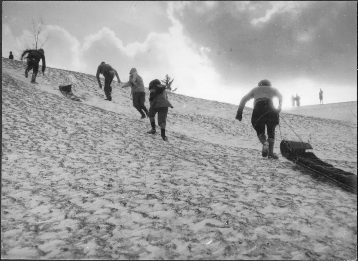 Black-and-white photograph of people climbing a snowy hill and pulling sleds