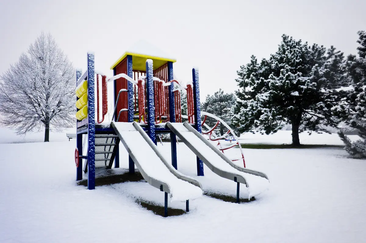 A snowy playground with snow piled on its slides and frosted trees behind