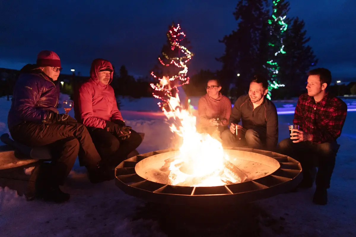 Five people sitting around a fire pit on a snowy evening holding drinks, with lit trees behind them