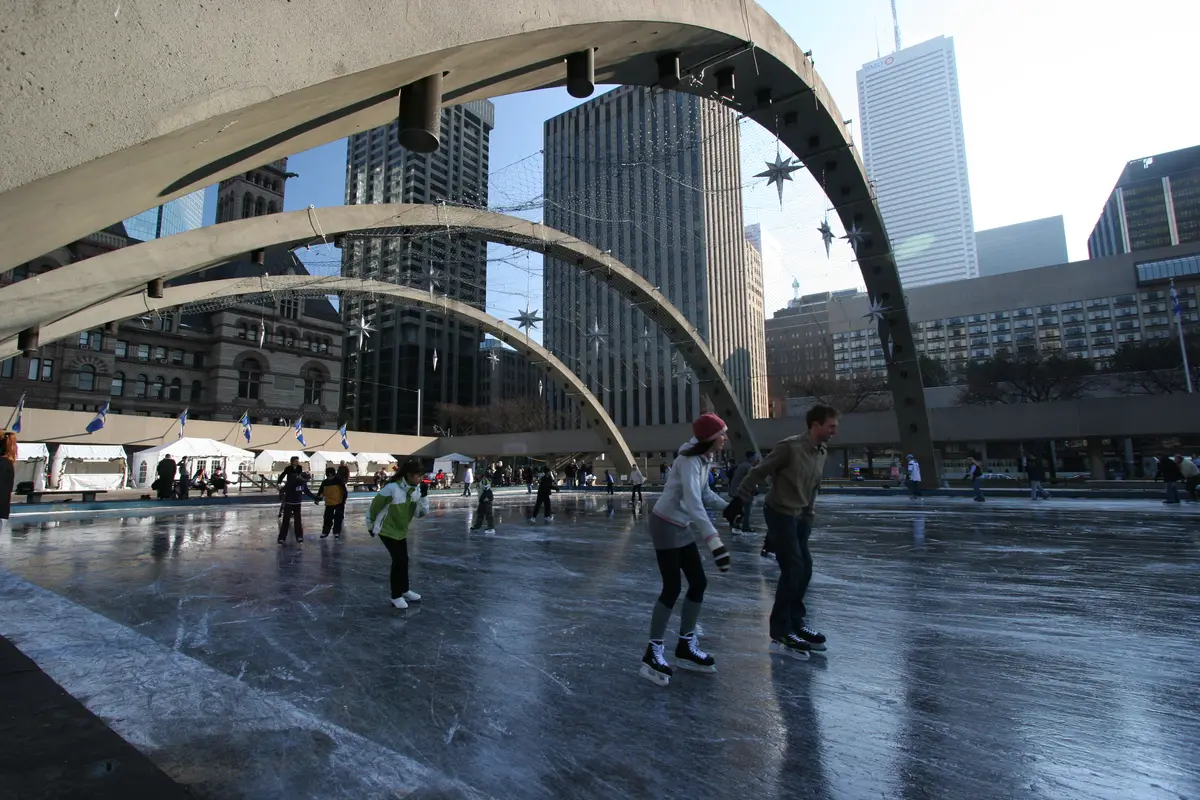 A couple skating side by side on the ice beneath concrete arches, with other skaters and city towers behind