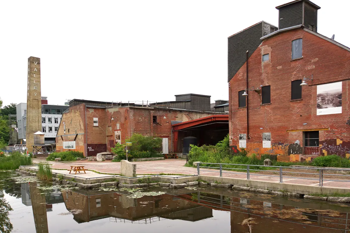 Red brick industrial buildings and a tall chimney of Evergreen Brick Works reflected in still water on a grey day