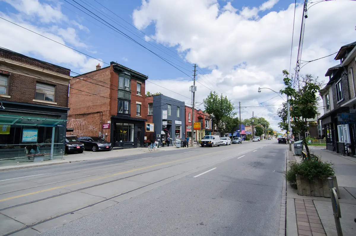 A sunny stretch of Queen Street East with brick storefronts, streetcar tracks and trees
