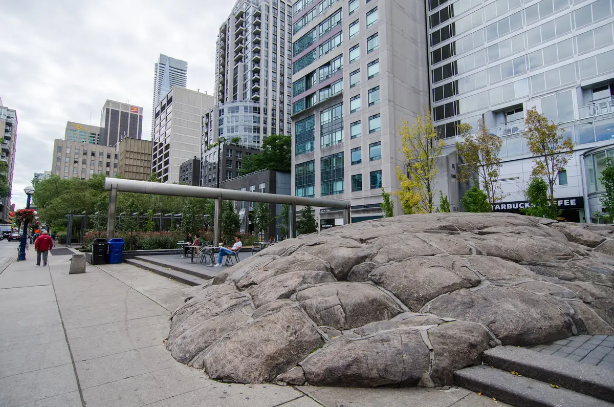 A large flat boulder in a paved plaza with benches, pine trees and apartment towers rising behind it.